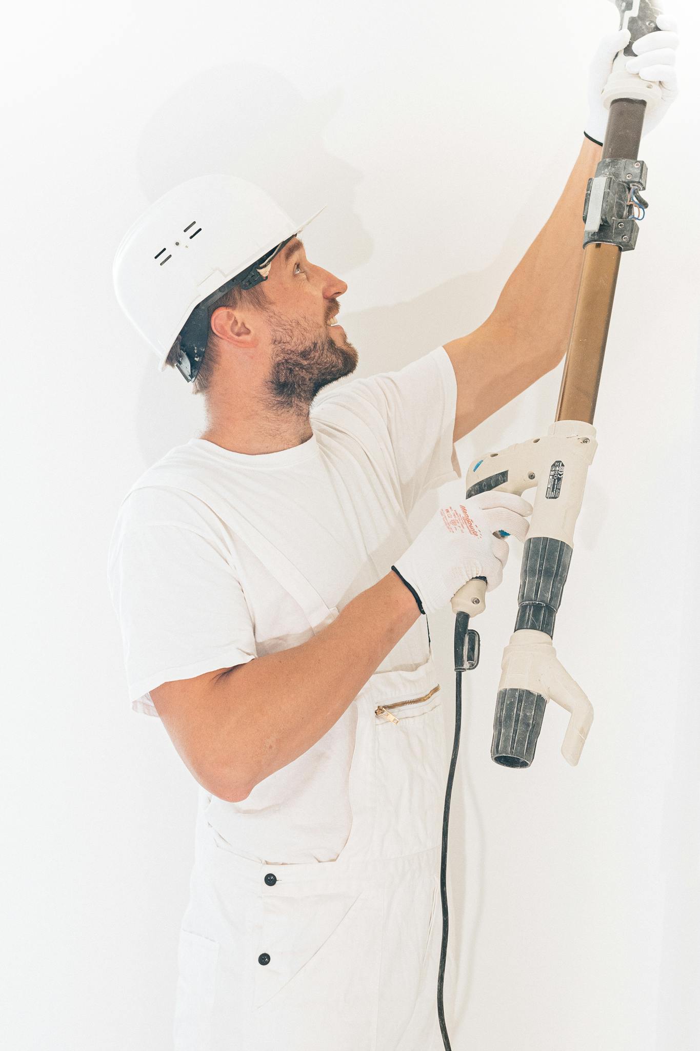 Mitarbeiter mit Schutzausrüstung verwendet einen Elektroschleifer an einer hellen weißen Wand im Innenbereich.